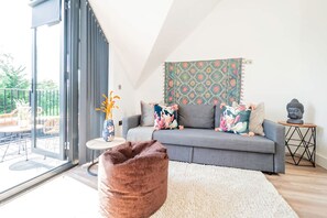 A modern living room with a grey sofa, colorful cushions, a patterned tapestry, a small round table, and a statue on a side table. - Omega Acton Top Floor (London)