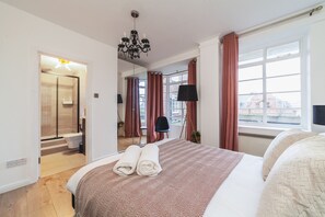 Ein Hotelzimmer mit einem großen Bett, einem Chandelier, einem Fenster mit Vorhängen, einem Badezimmer mit Dusche und Blick auf die Stadt.