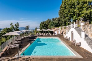 A rectangular pool with a wooden deck, surrounded by lounge chairs and umbrellas, with a stone staircase and a view of the landscape. - Masaccio Apartament in Villa Guinigi (Capannori)