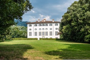 A large white building with multiple windows and a central entrance, surrounded by a well-maintained lawn and dense trees. - Masaccio Apartament in Villa Guinigi (Capannori)