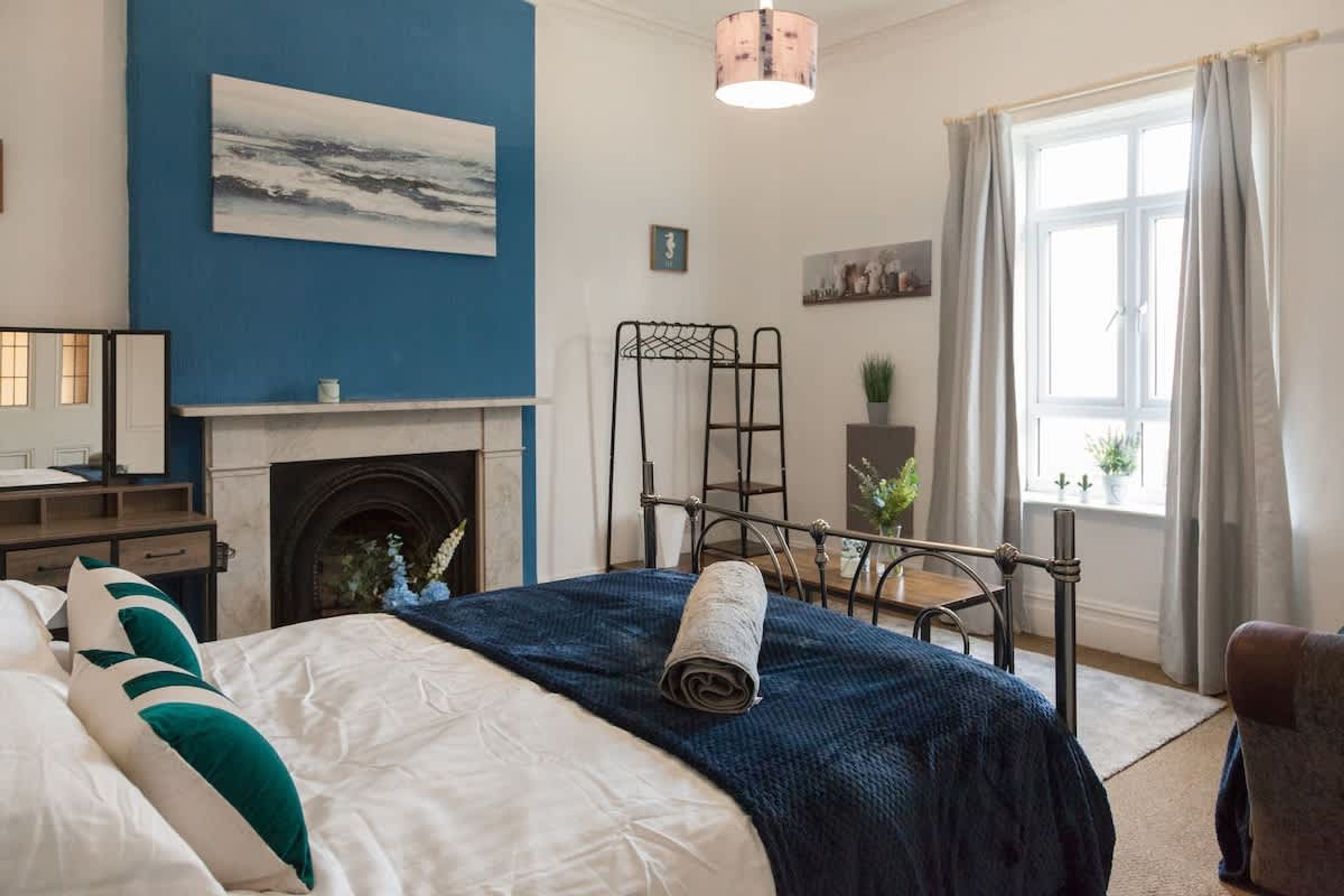 A bedroom with a bed, a fireplace, a window with curtains, a desk with a lamp, and a wall-mounted picture.