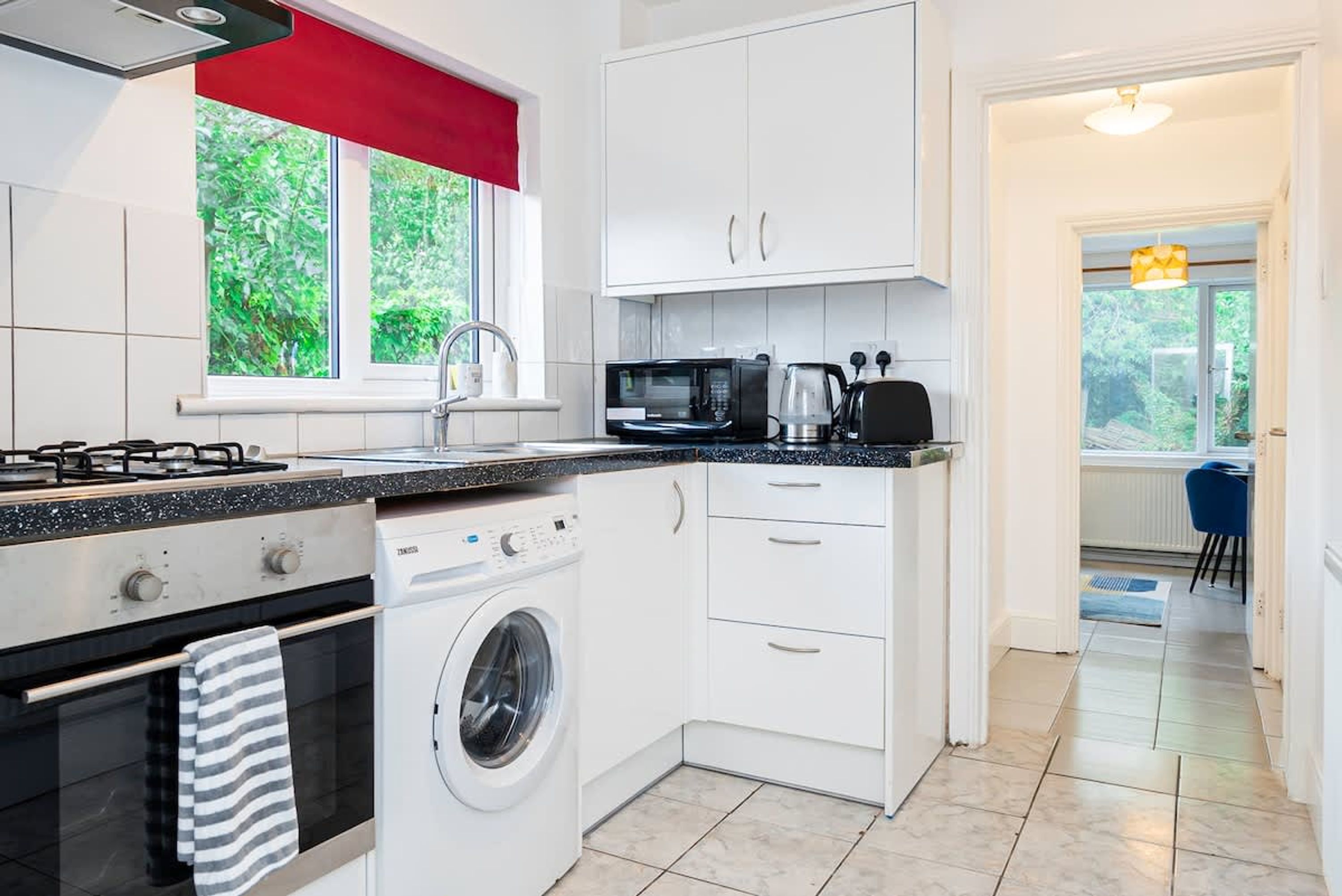 Une cuisine compacte équipée d’une cuisinière à gaz, d’un four à micro-ondes, d’un lave-linge et de meubles de rangement blancs.