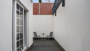 En smal udendørs terrasse med træterrasse, to stole, et lille rundt bord og en hvid væg med et træhegn overfor.