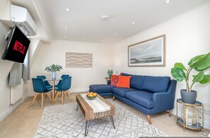 Ein Wohnzimmer mit einem blauen Sofa, einem Couchtisch mit Obst, einem Fernseher, der 'NETFLIX' zeigt, und einem großen gerahmten Bild an der Wand.
