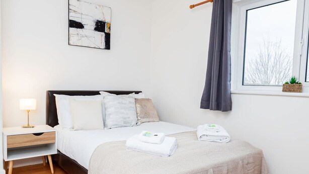 Un dormitorio con cama, mesita de noche, lámpara y ventana con cortinas.