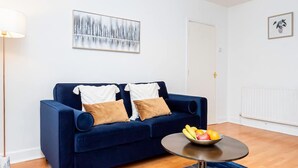 Un sofá azul con dos almohadas, una mesa de centro redonda con fruta, una lámpara de pie y un cuadro en la pared.