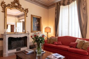 Ein Zimmer mit Kamin, einem roten Sofa, einer Blumenvase, einem Spiegel und einem Gemälde an der Wand.