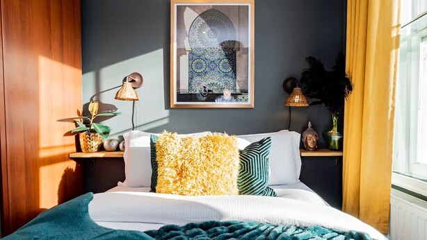 Una camera da letto con un letto, cuscini, una lampada, un quadro appeso al muro e una finestra con tende.