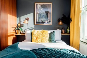 A bedroom with a bed, pillows, a lamp, a wall-mounted picture, and a window with curtains. - Stylish 2BR Near Hyde Park Museums Kensington (London)