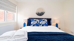 Ein ordentlich bezogenes Bett mit einem blau-weißen Kissen, einer blauen Decke und einem Stapel weißer Handtücher.