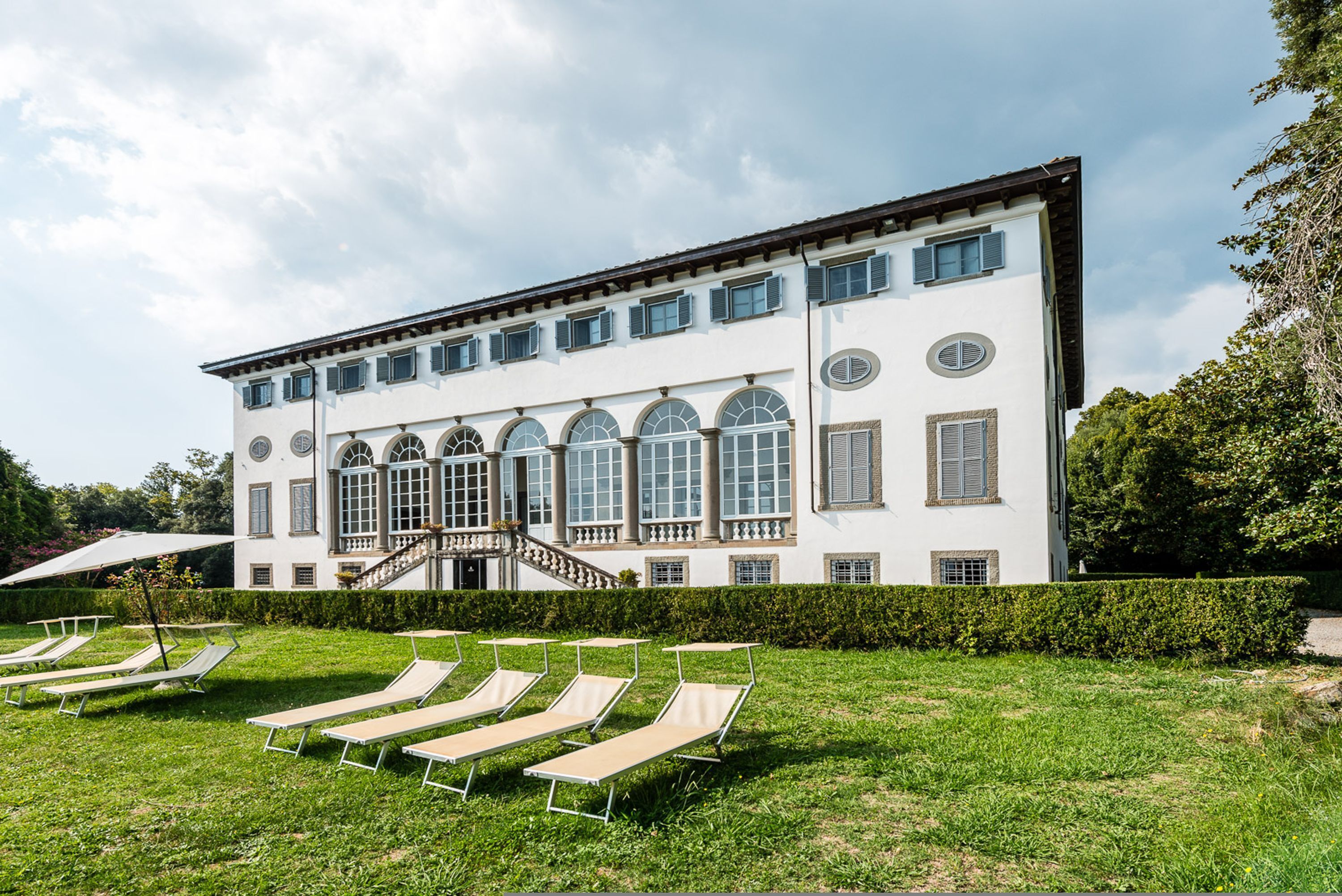 A large white building with multiple windows and a staircase leading to a balcony. There are several lounge chairs arranged in the foreground.