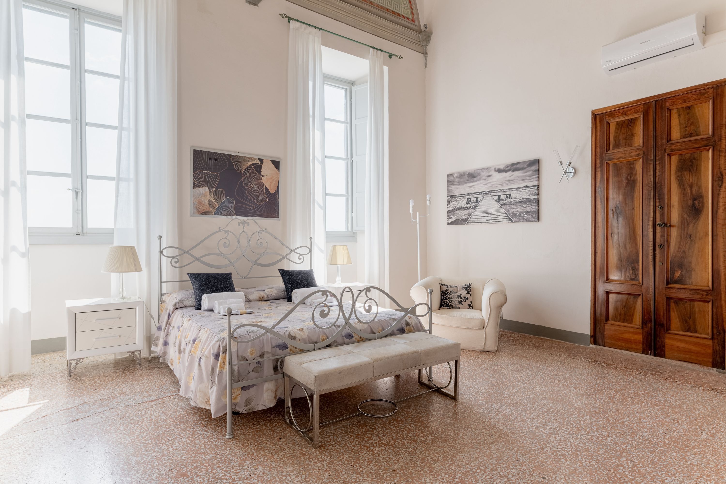 A bedroom with a bed, bench, bedside table, lamp, and artwork on the walls.