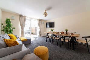 A modern living room with a sofa, ottoman, dining table, chairs, and a TV. - Lovely House in Stratford (London)