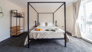 En himmelseng med hvid dyne, et natbord med en skål croissanter og en lille hylde med en plante.