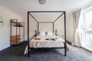 A four-poster bed with a white bedspread, a bedside table with a bowl of croissants, and a small shelf with a plant. - Lovely House in Stratford (London)
