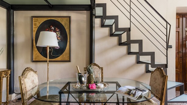 Un comedor con una mesa de vidrio, sillas, un cuadro enmarcado y una escalera.
