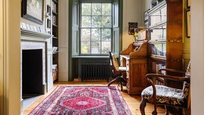 Ein Zimmer mit Kamin, großem Fenster, Holzstuhl, Bücherregal und Teppich mit Muster.