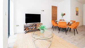 A modern living room with a flat-screen TV, a dining area with orange chairs, and a round coffee table with a plant. - Modern Luxurious Retreat in Acton w Great Links (London)