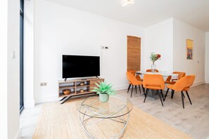 A modern living room with a flat-screen TV, a dining area with orange chairs, and a round coffee table with a plant. - Modern Luxurious Retreat in Acton w Great Links (London)