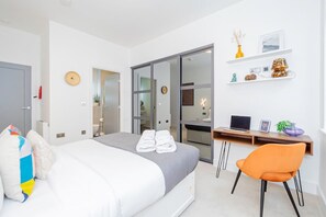 Et moderne hotelværelse med seng, skrivebord, stol, garderobeskab og et badeværelse, der kan ses igennem en glasdør.