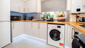 A compact kitchen with white cabinets, a microwave, a washing machine, and a window with a view of greenery. - Spacious 2BR Flat Summertown Sleeps 7 5 Star (Oxford)