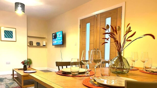 A dining area with a wooden table set for four, a TV mounted on the wall, and a door leading to another room. - Spacious 2BR Flat Summertown Sleeps 7 5 Star (Oxford)