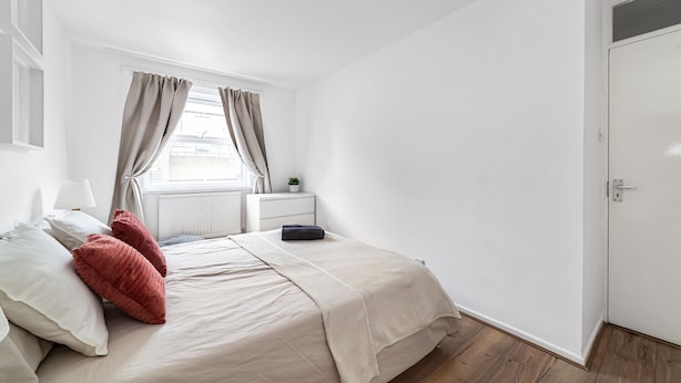 A bedroom with a bed, pillows, a nightstand, a window with curtains, a door, and a radiator. - 3 Bed House With Garden in London Fields (London)