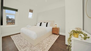 Quarto com cama, mesa de cabeceira, abajur e vista para edifícios pela janela.