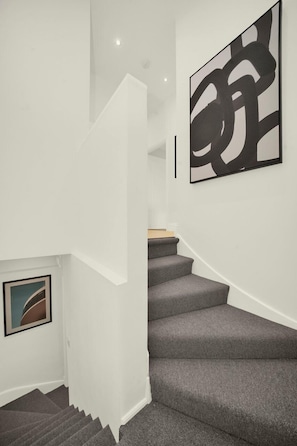 Una escalera con peldaños cubiertos de moqueta gris, paredes blancas y un cuadro abstracto de gran tamaño a la derecha.