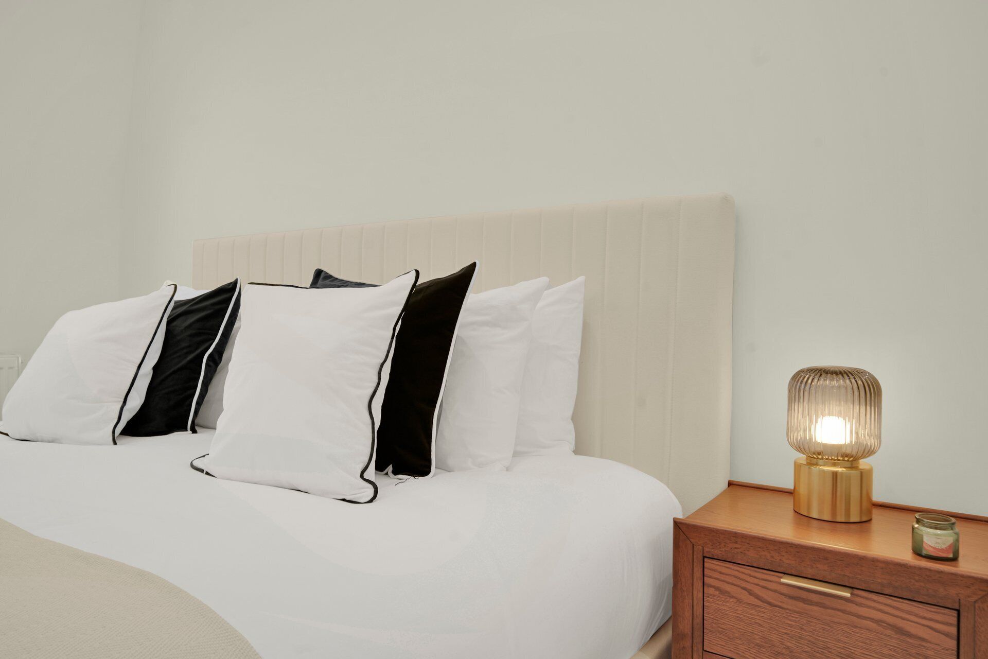 A neatly made bed with white and black pillows, a wooden nightstand with a lamp, and a small jar.
