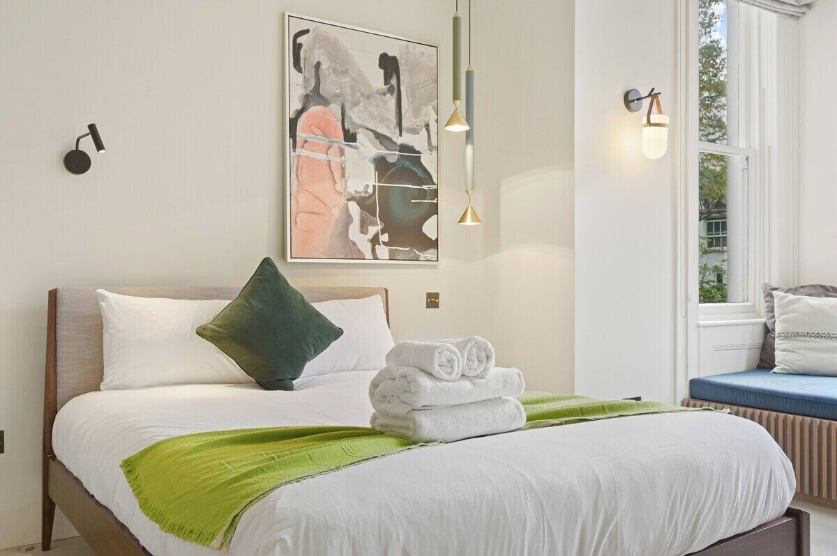 A neatly made bed with white linens and a green throw, a wall-mounted abstract painting, and a window with a view of greenery.