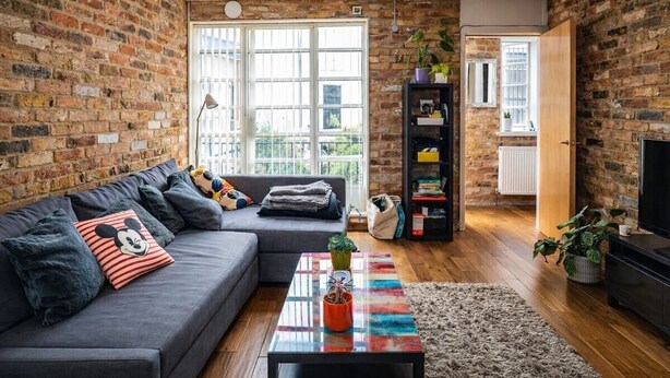 Ein Wohnzimmer mit Backsteinwand, einem grauen Sofa, einem bunten Couchtisch und einem Fernsehschrank.
