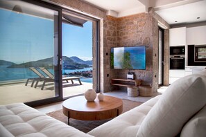 A modern living room with a large TV, a stone accent wall, and a balcony with a view of the sea. - Smirties Villa Phaena 2bed (Lefkada)