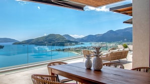 A wooden table with wicker chairs, two pineapple-shaped vases, and a glass railing overlooking a coastal view with mountains. - Smirties Villa Phaena 2bed (Lefkada)
