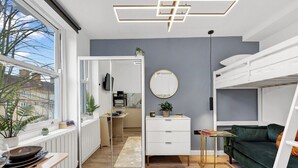 A modern interior with a kitchen, dining area, and living space, featuring a bunk bed, a round mirror, and a view through a glass door. - Single Studio Flat close to Camden! (London)