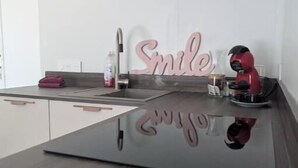 A modern kitchen with a dark countertop, a sink, a faucet, a pink 'Smile' sign, and a red helmet on the counter. - Bright 2-Bedroom Apartment in the Heart of the Vineyards – 25 Minutes from Bordeaux (Prignac-et-Marcamps)