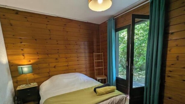 A wooden-paneled room with a bed, a nightstand, a lamp, and a window with curtains. - Bright 2-Bedroom Apartment in the Heart of the Vineyards – 25 Minutes from Bordeaux (Prignac-et-Marcamps)