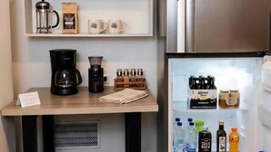 Une petite cuisine équipée d’une machine à café, d’un moulin à café, d’un réfrigérateur rempli de boissons et d’une étagère contenant des paquets de café et des tasses.