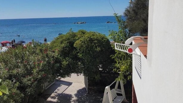 Vista del mar desde un balcón con barandilla blanca, un pequeño jardín con una banca y un cielo azul despejado.