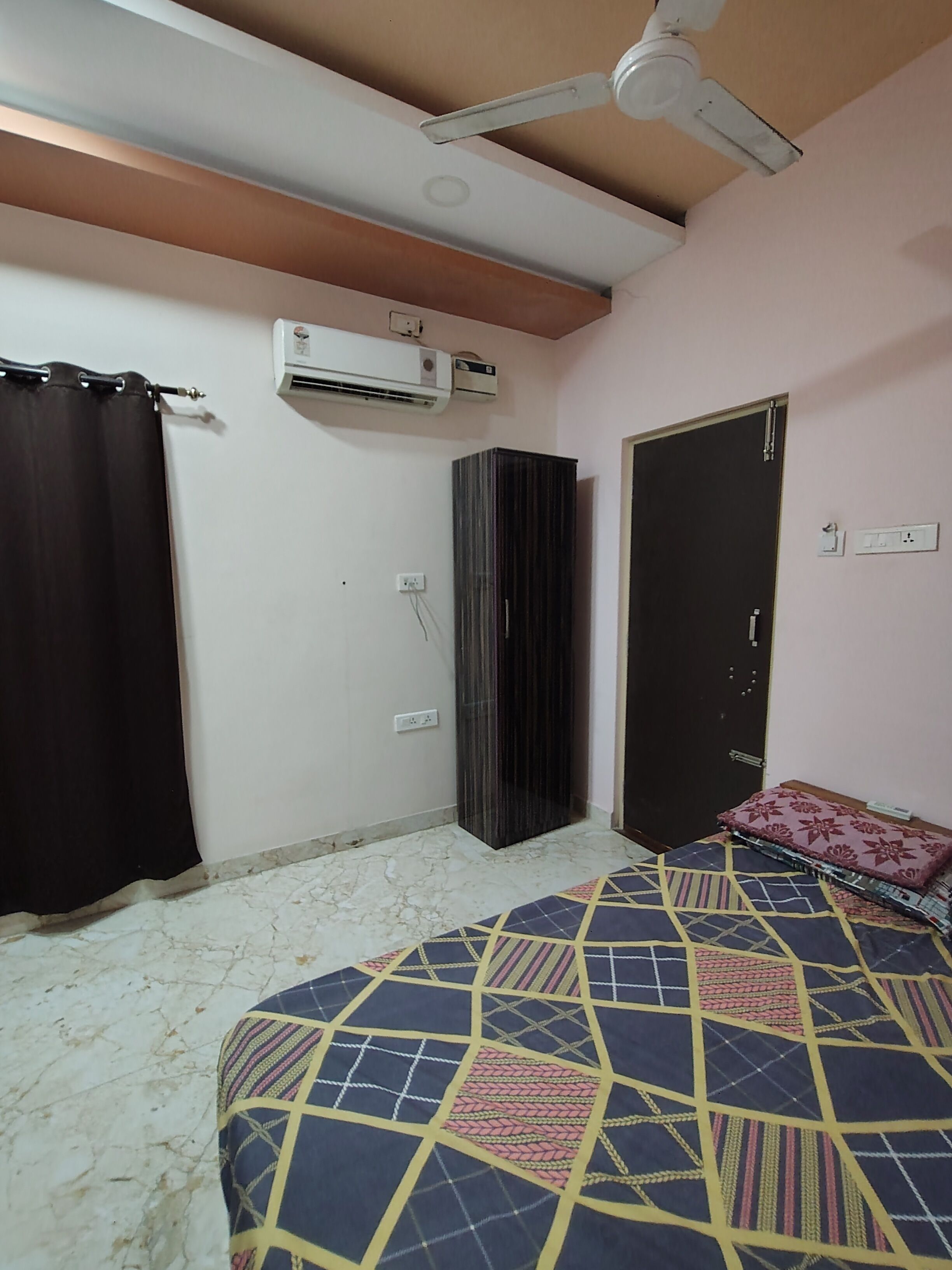 A bedroom with a bed, a ceiling fan, an air conditioning unit, a wardrobe, and a door.
