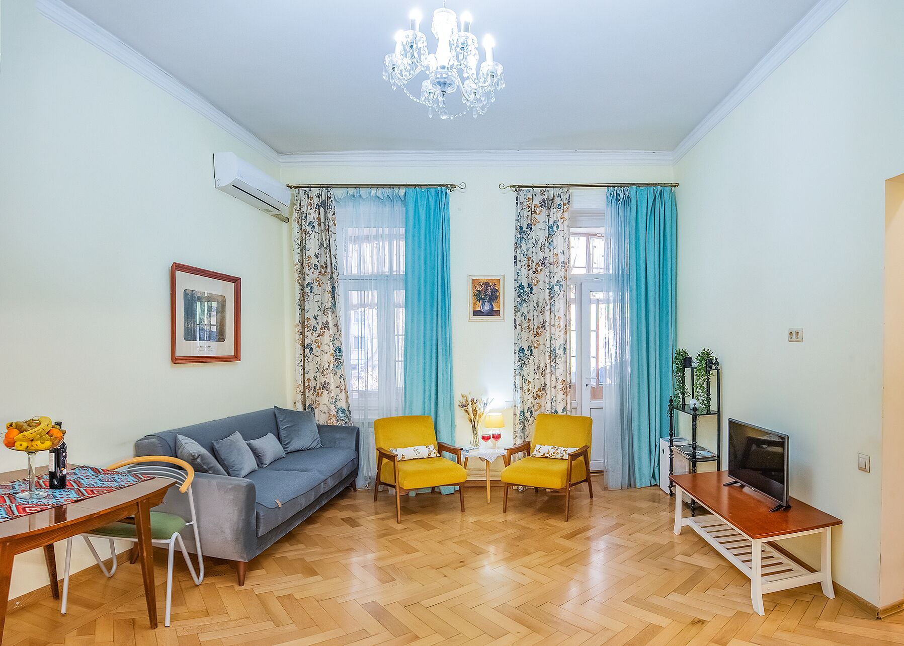 A living room with a sofa, two yellow chairs, a TV on a stand, and a chandelier.