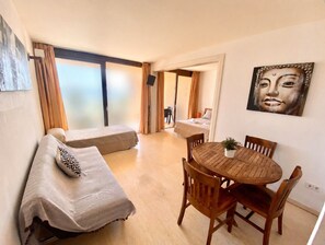 A room with a bed, a dining table with chairs, a painting on the wall, and a large window with curtains.
