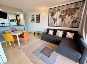 A living room with a sofa, a dining table with chairs, a kitchen area, and a world map artwork on the wall.