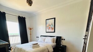 Un dormitorio con cama, mesita de noche, ventilador de techo, ventana con cortinas, un cuadro en la pared y un tapete estampado.