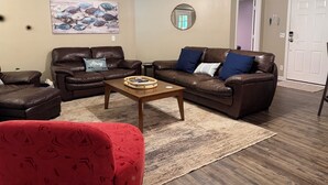 Ein Wohnzimmer mit zwei braunen Ledersofas, einem rot gemusterten Sessel, einem Holztisch, einem Bild von Fischen, einem Spiegel und einer weißen Tür.