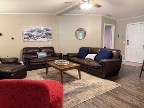Ein Wohnzimmer mit zwei braunen Ledersofas, einem rot gemusterten Sessel, einem Holztisch, einem Bild von Fischen, einem Spiegel und einer weißen Tür.