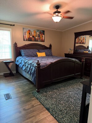 A bedroom with a bed, a ceiling fan, a painting, a dresser, and a window.