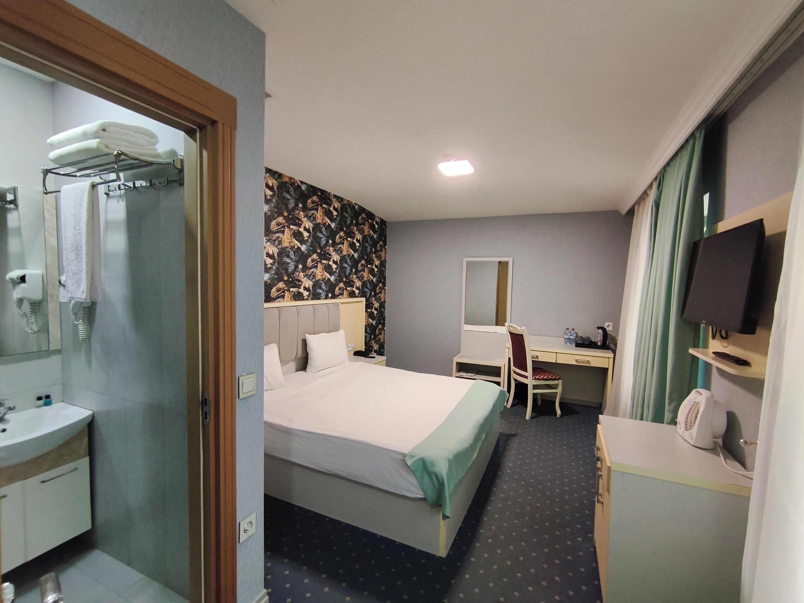 A hotel room with a bed, a desk, a chair, a TV, a mirror, and a bathroom with a sink and towel rack.