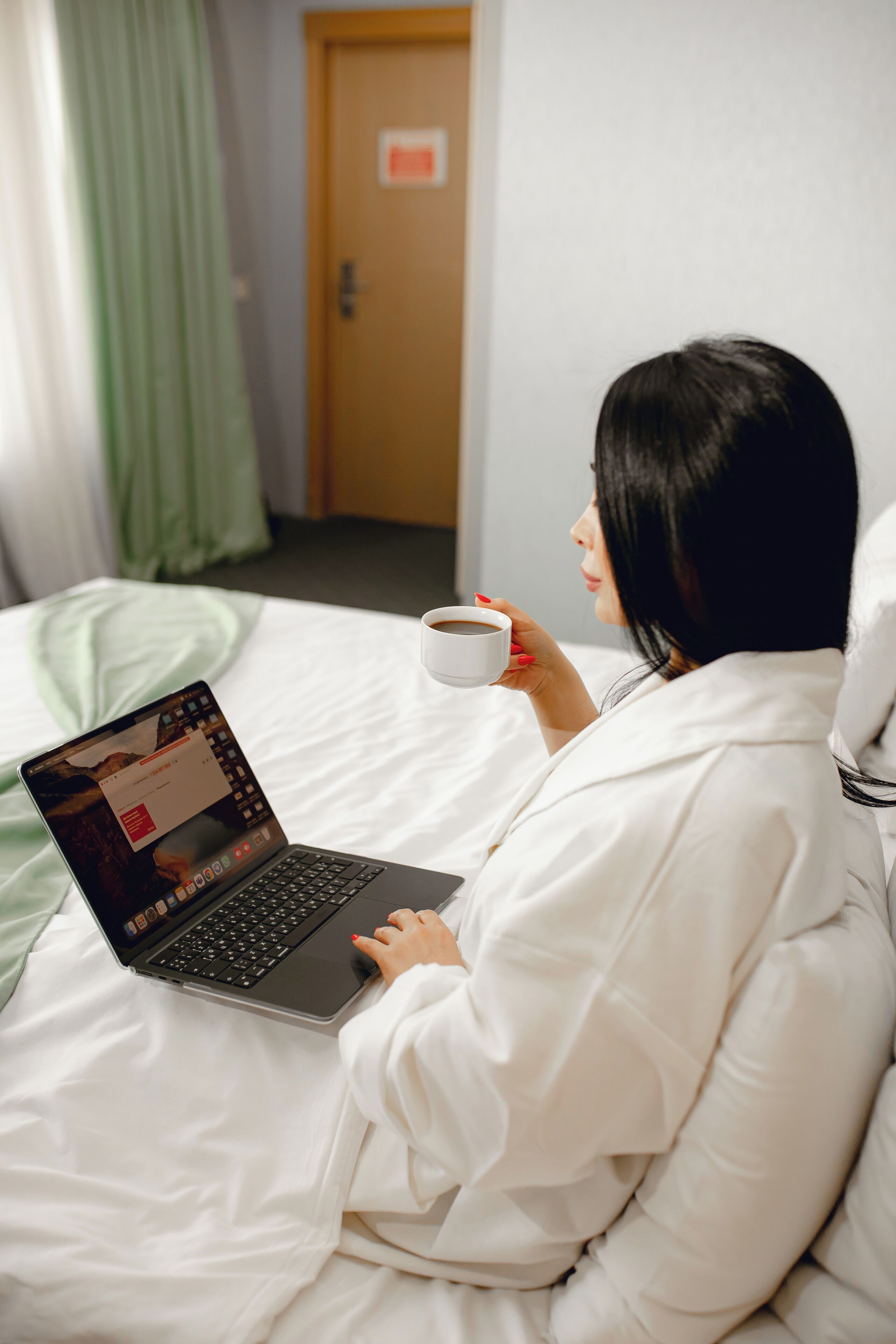 Iemand zit op een bed met een laptop en een kopje, in een kamer met een deur en gordijnen.