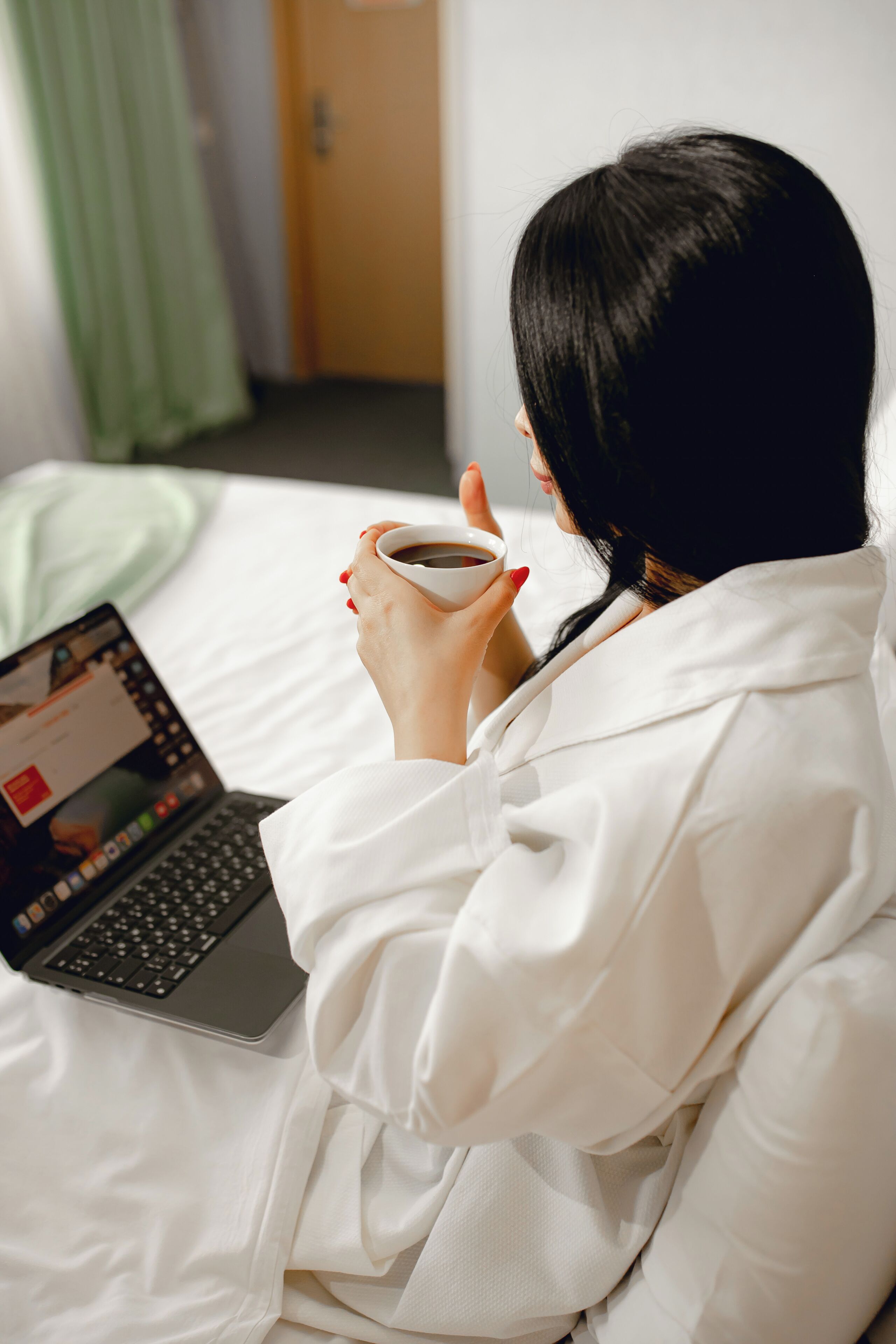 Una persona sentada en una cama con una computadora portátil y sosteniendo una taza.